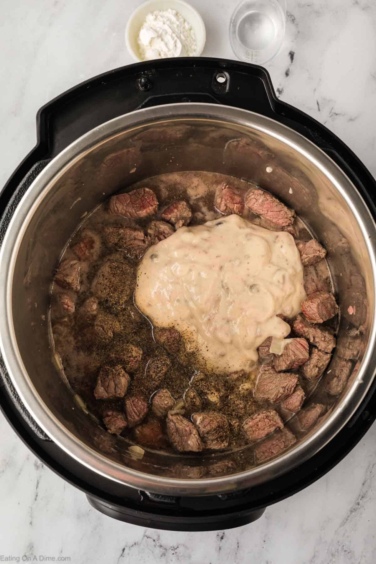 An Instant Pot brimming with beef tips bathed in a creamy mushroom gravy, while a small bowl of flour rests nearby on the marble countertop.