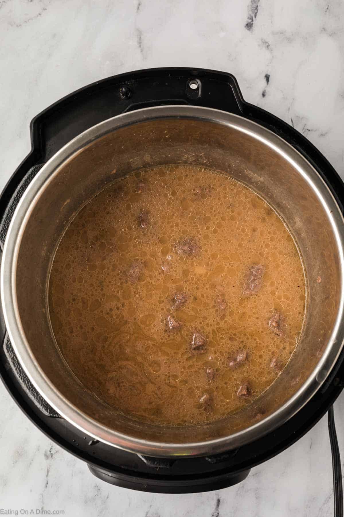 A top view of an Instant Pot on a marble countertop reveals a bubbling brown stew with chunks of beef tips, simmering to perfection in its savory gravy.