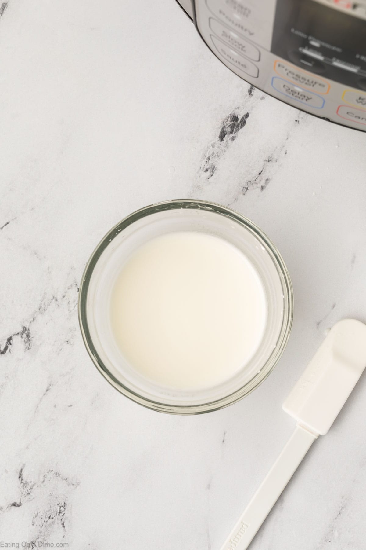 A small glass bowl filled with a white liquid sits on a marble countertop next to a white spatula, hinting at the delicious Instant Pot Beef Tips and Gravy being prepared. Part of an appliance is visible in the corner, ready to work its magic.