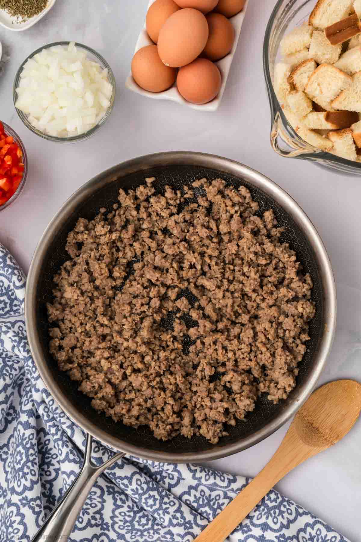 Cooked sausage crumbles in a large skillet with eggs on a plate with bread cubes and diced onions on the side