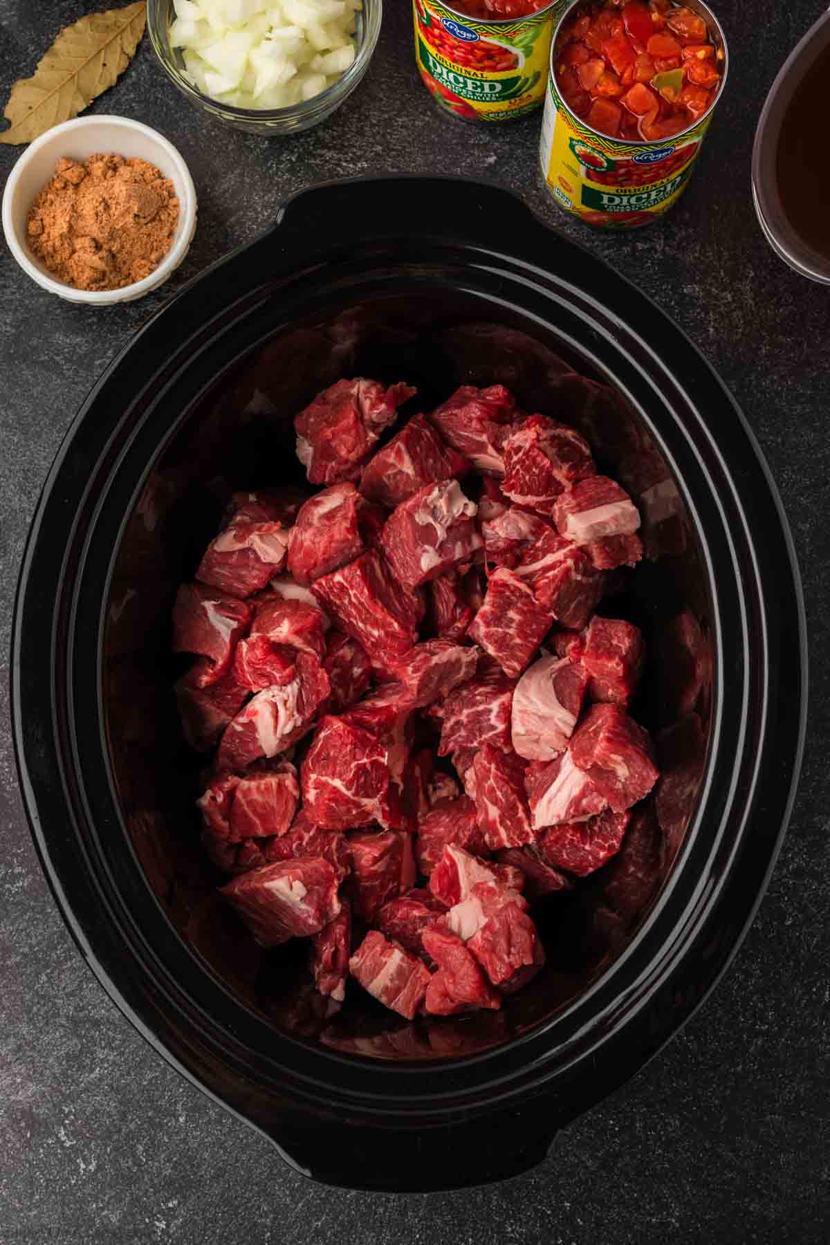 A slow cooker filled with uncooked chunks of beef awaits transformation into a savory Mexican beef stew. Nearby are small bowls of diced onions and brown spice mix, plus cans of diced tomatoes and a cup of broth. A bay leaf is placed on the table, ready to infuse its flavor.