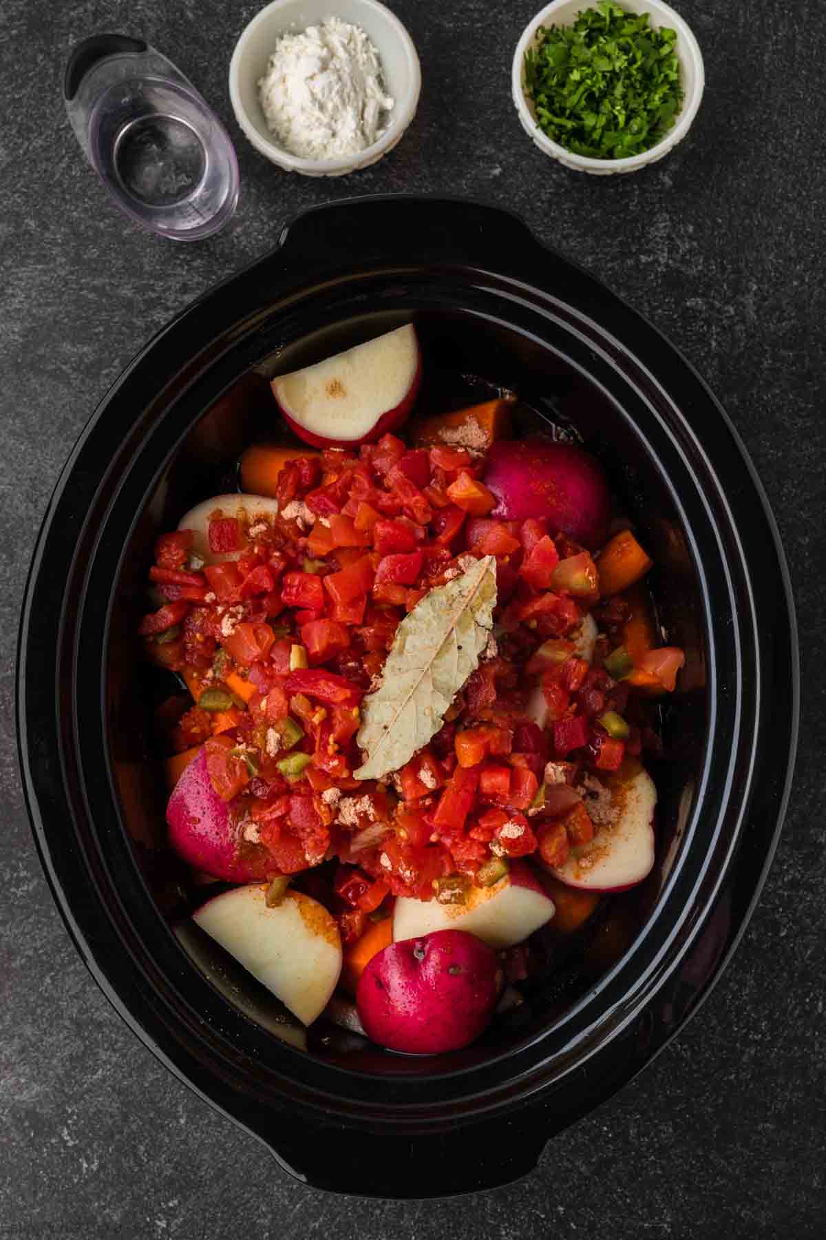 An overhead view of a slow cooker filled with diced tomatoes, red potatoes, and a bay leaf hints at the rich flavors of a Mexican beef stew. Small bowls with flour, chopped herbs, and a cup of water are placed beside it on a dark surface.