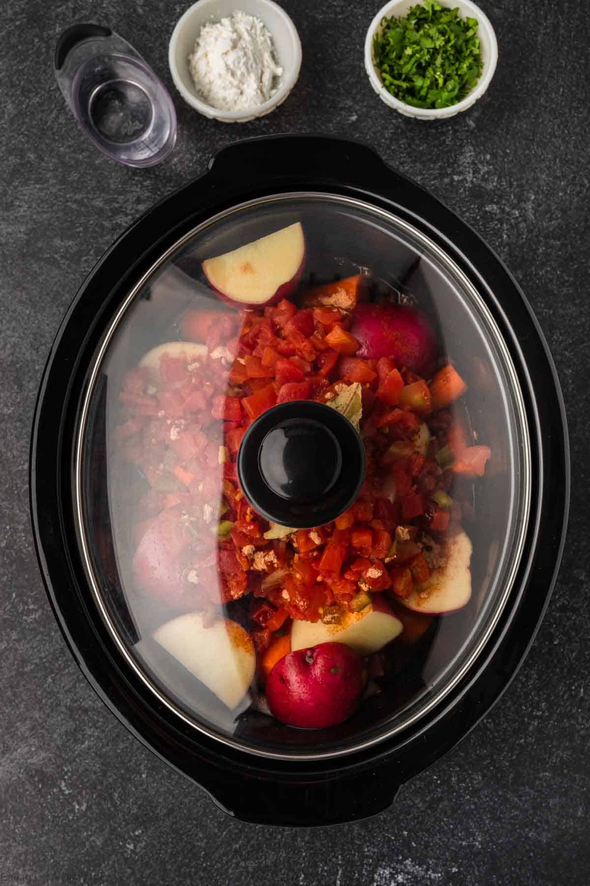 A slow cooker filled with a hearty Slow Cooker Mexican Beef Stew sits on a dark countertop. Chopped tomatoes, potatoes, and spices mingle within, promising rich flavors. Nearby are small bowls of flour and chopped herbs, alongside a small glass of water. The slow cooker lid is in place.