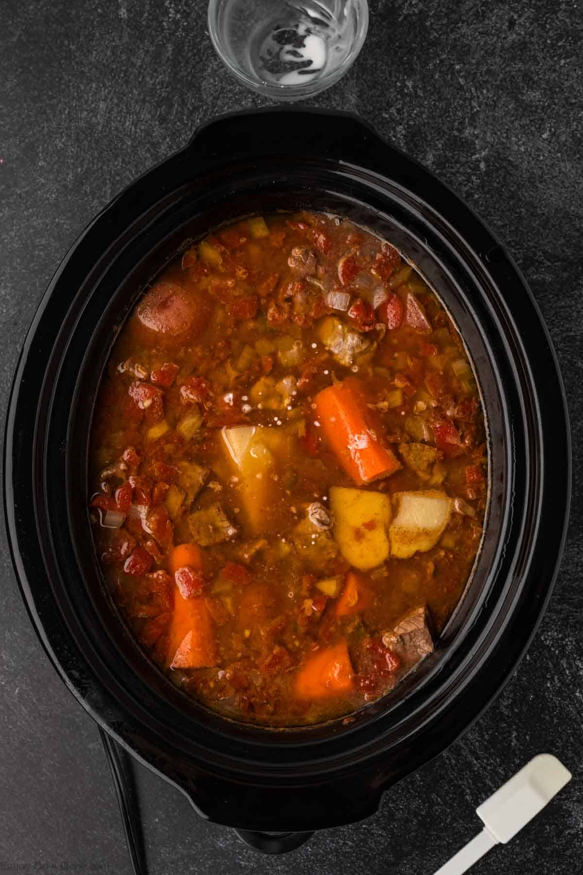 A slow cooker brimming with Mexican beef stew features chunks of beef, potatoes, carrots, tomatoes, and onions in a rich broth. A glass bowl and a white spatula are beside this enticing creation on a dark countertop.