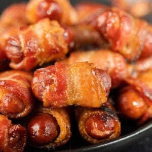 Close-up of a plate of bacon-wrapped little smokies. The sausages are tightly wrapped with crispy, golden-brown bacon strips, giving a savory and appetizing appearance. The dish is presented in a way that highlights the texture and juiciness of the bacon and sausages.