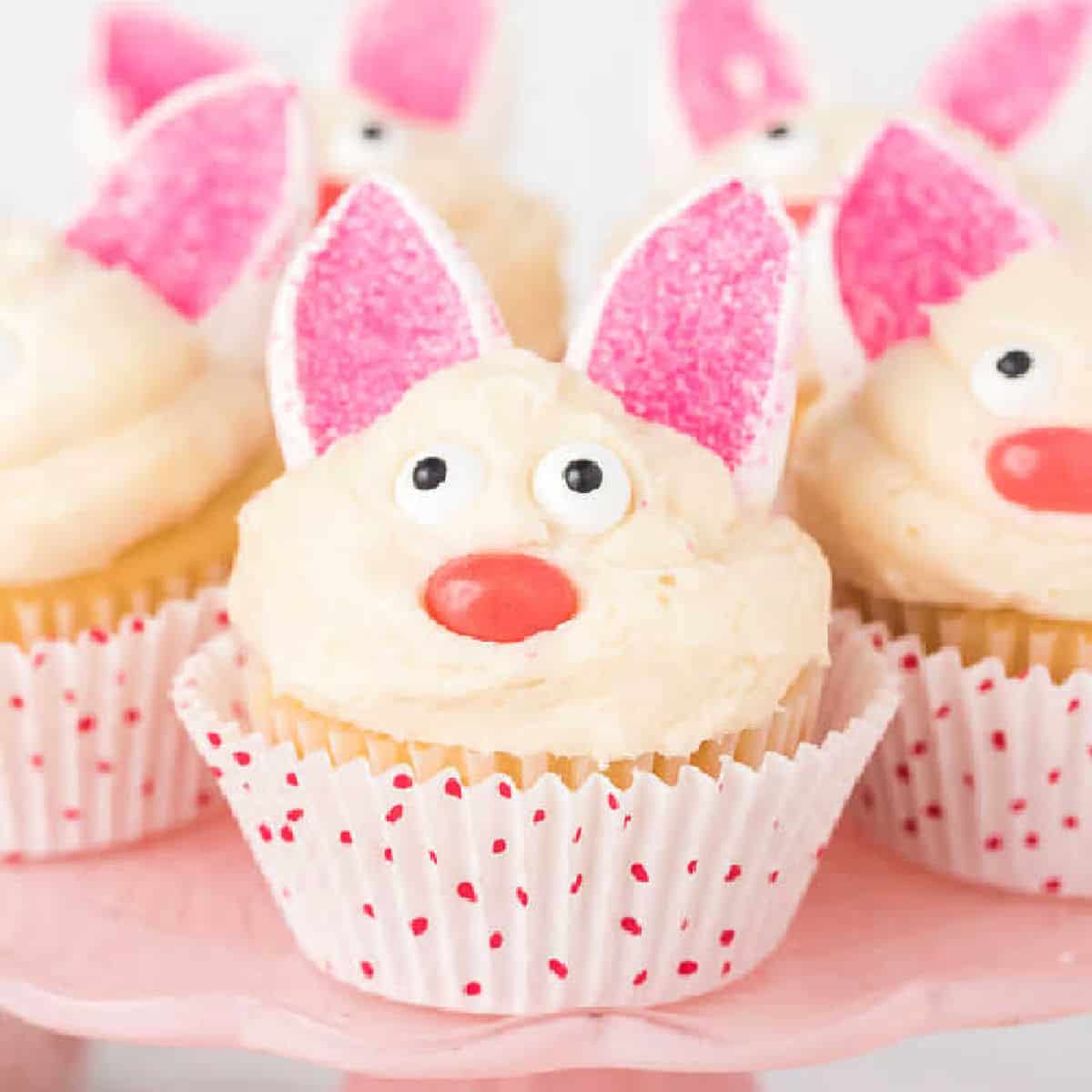 Easter Bunny Cupcakes decorated to look like bunny faces, with pink sugar-coated ears, white frosting, candy eyes, and red noses, displayed on a pink cake stand. The cupcakes are in white wrappers with red polka dots.