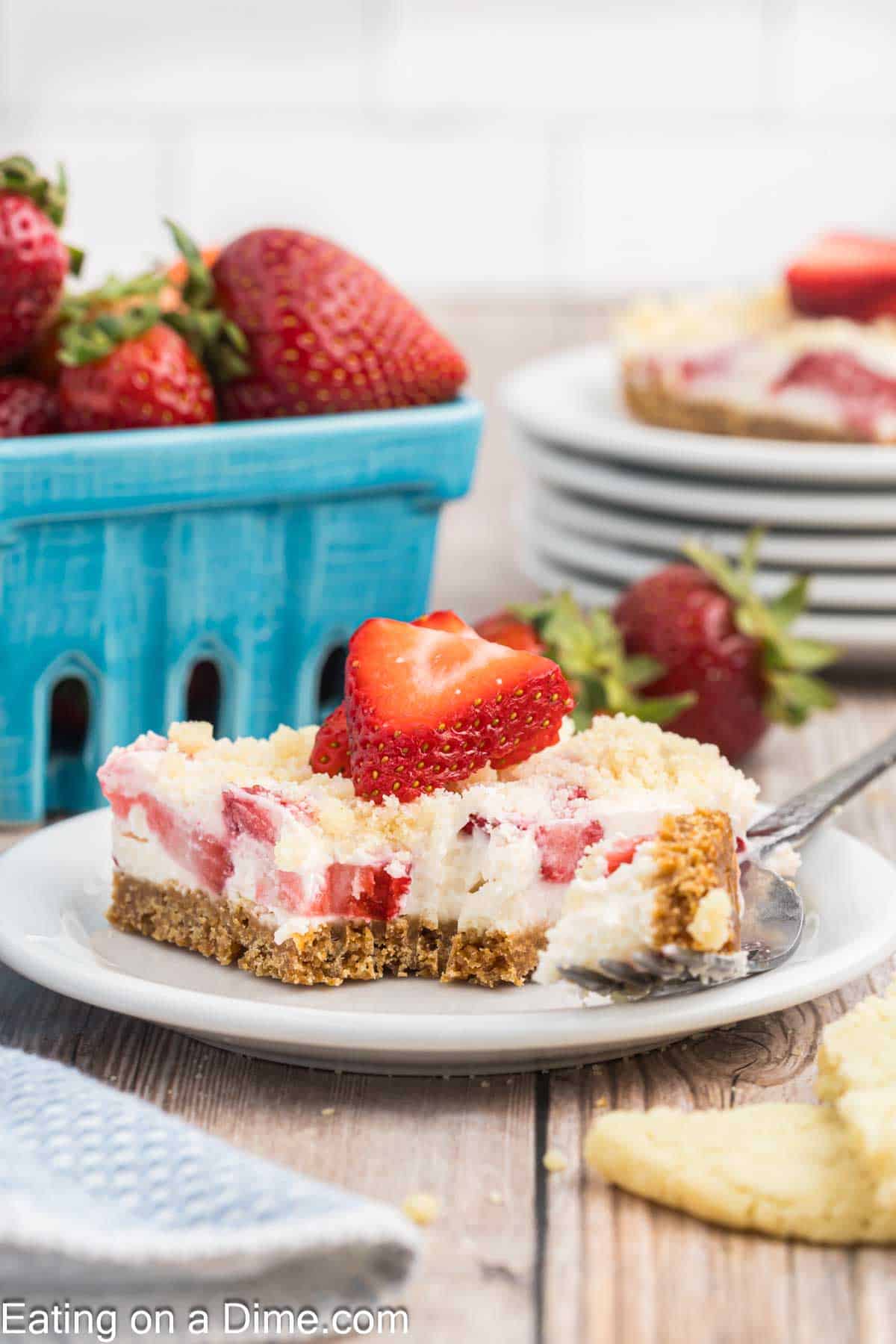 A close-up of a slice of strawberry cheesecake on a white plate with a fork. The cheesecake has a graham cracker crust and fresh strawberries on top, reminiscent of an indulgent strawberry crunch bars recipe. Behind the plate, there's a container of strawberries and a stack of plates with more cheesecake slices.
