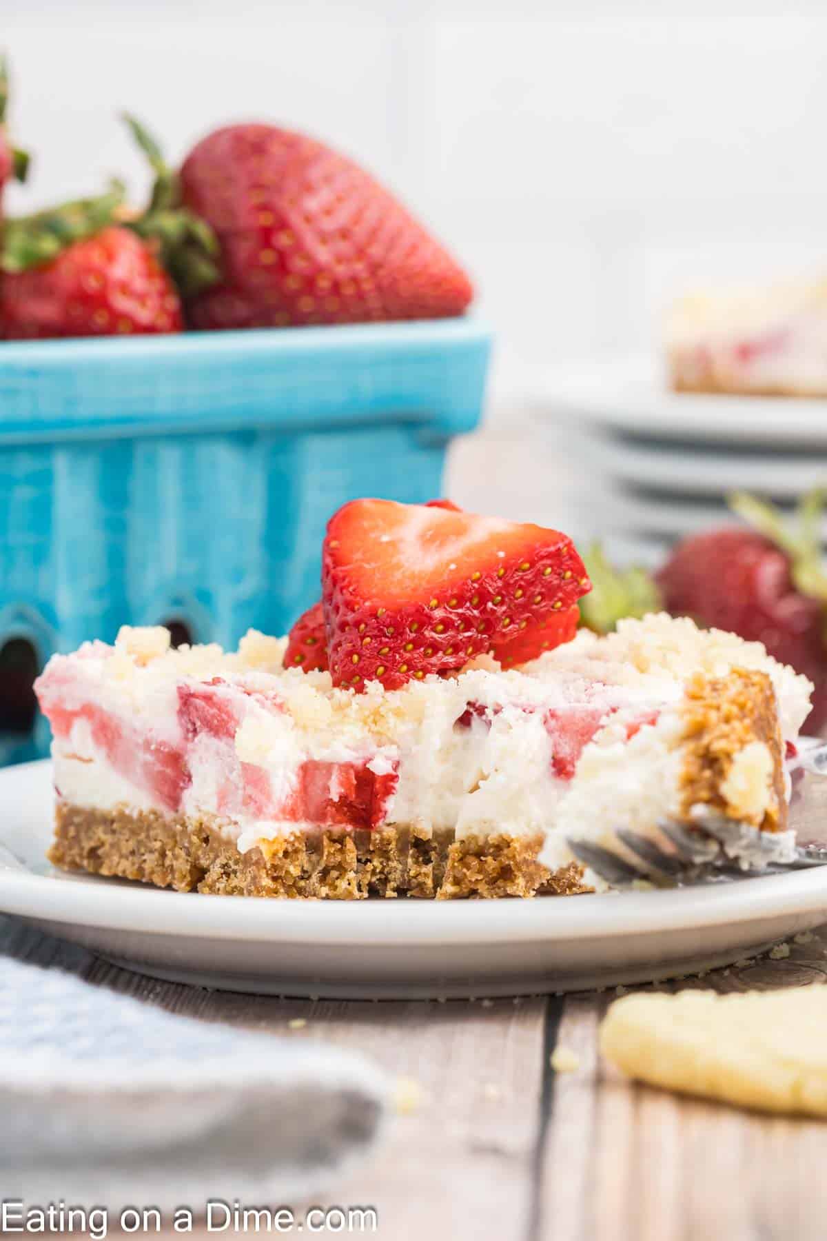 A slice of creamy strawberry cheesecake with a graham cracker crust sits on a white plate. A fork with a bite of the cheesecake rests beside it. Fresh strawberries garnish the slice, evoking memories of a delightful strawberry crunch bars recipe, with a basket of strawberries in the background on a wooden table.
