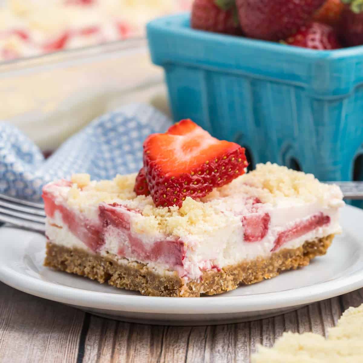 A slice of strawberry crunch bars sits on a white plate. The dessert consists of a crumbly base, creamy filling with strawberry chunks, and a crumb topping. A halved strawberry garnishes the top. A basket of fresh strawberries is visible in the background, enticing you to try this delicious recipe.