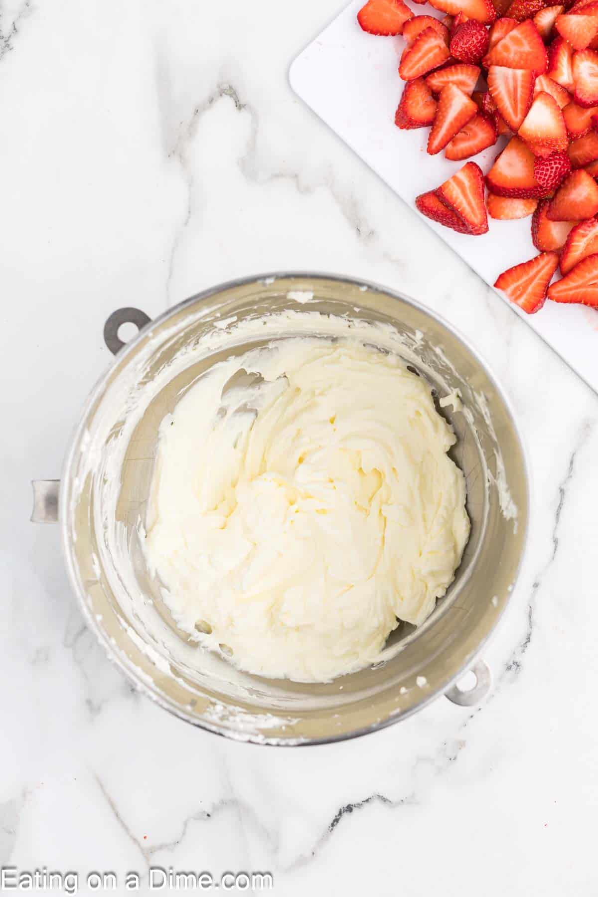 A mixing bowl filled with whipped cream sits on a white marble countertop. To the upper right, there is a cutting board with sliced strawberries, perfect for your strawberry crunch bars recipe. The background is clean and bright.