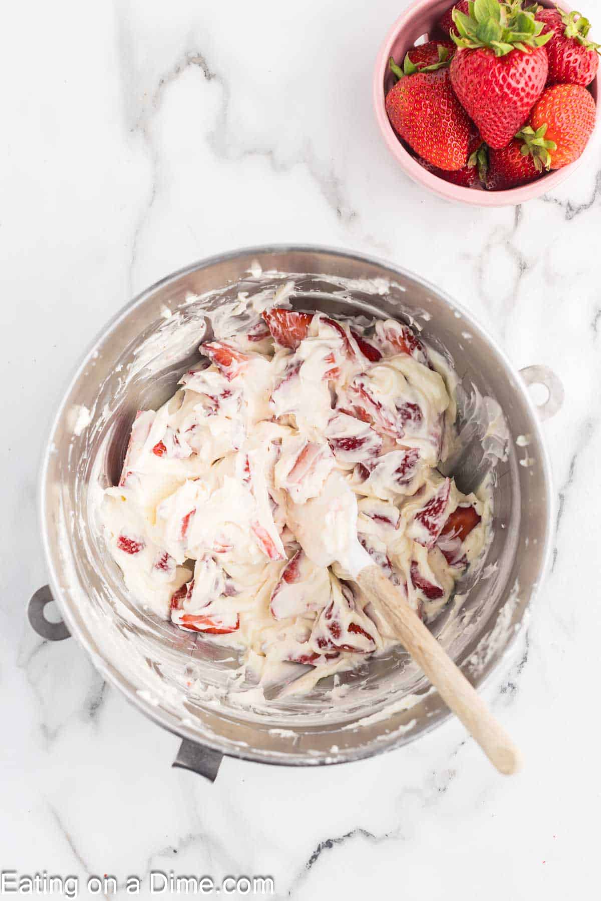 A mixing bowl filled with cream-covered strawberry slices and a wooden spoon sits on a white marble surface, resembling the start of a delightful strawberry crunch bars recipe. Next to the bowl, there's a small pink bowl containing whole fresh strawberries.