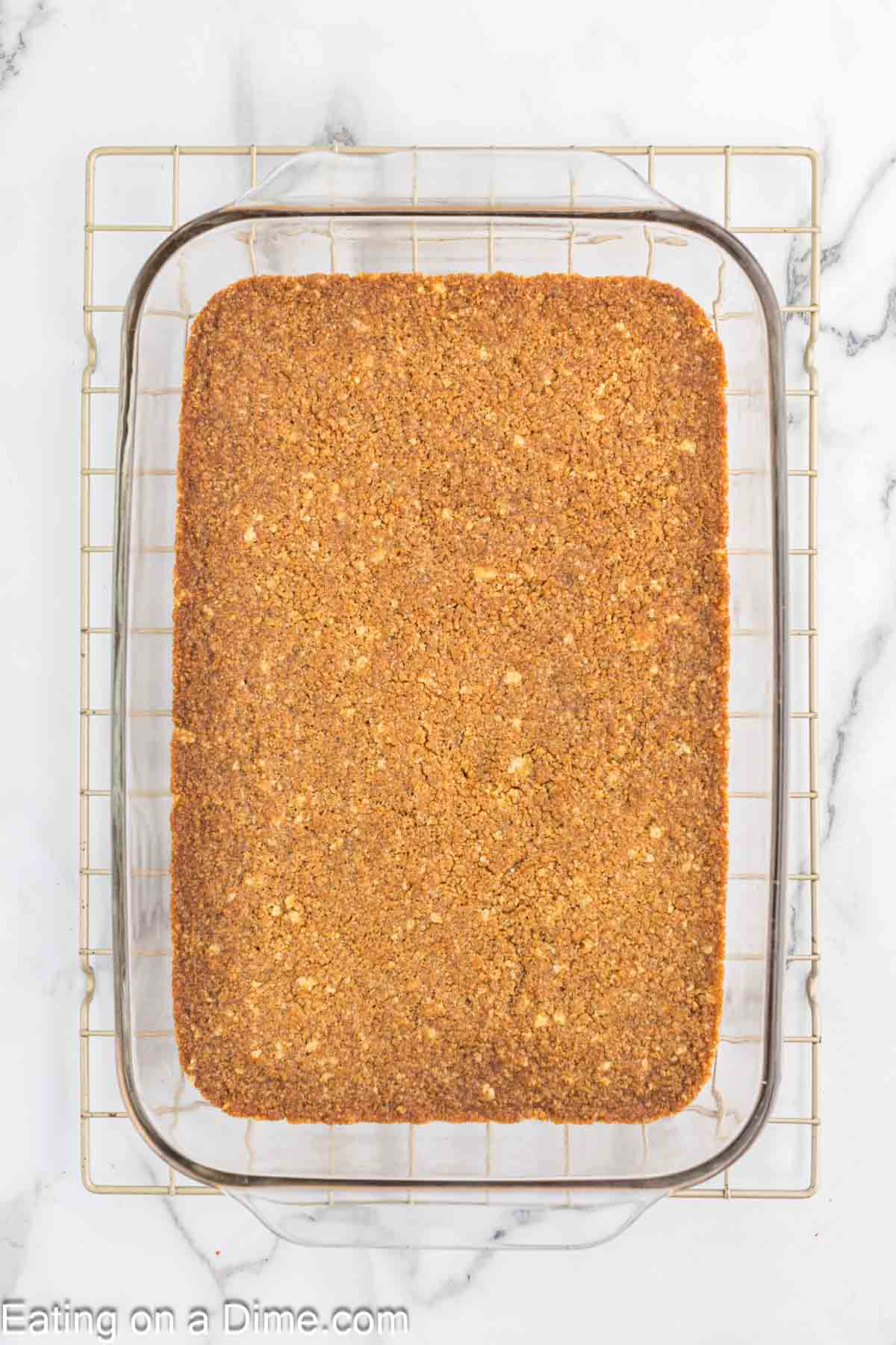 A large glass baking dish filled with evenly browned, baked oatmeal sits on a wire cooling rack atop a white marble surface. The dish's contents have a crumbly texture and are slightly cracked on the surface. The text "Eating on a Dime.com" is visible at the bottom, featuring the strawberry crunch bars recipe.