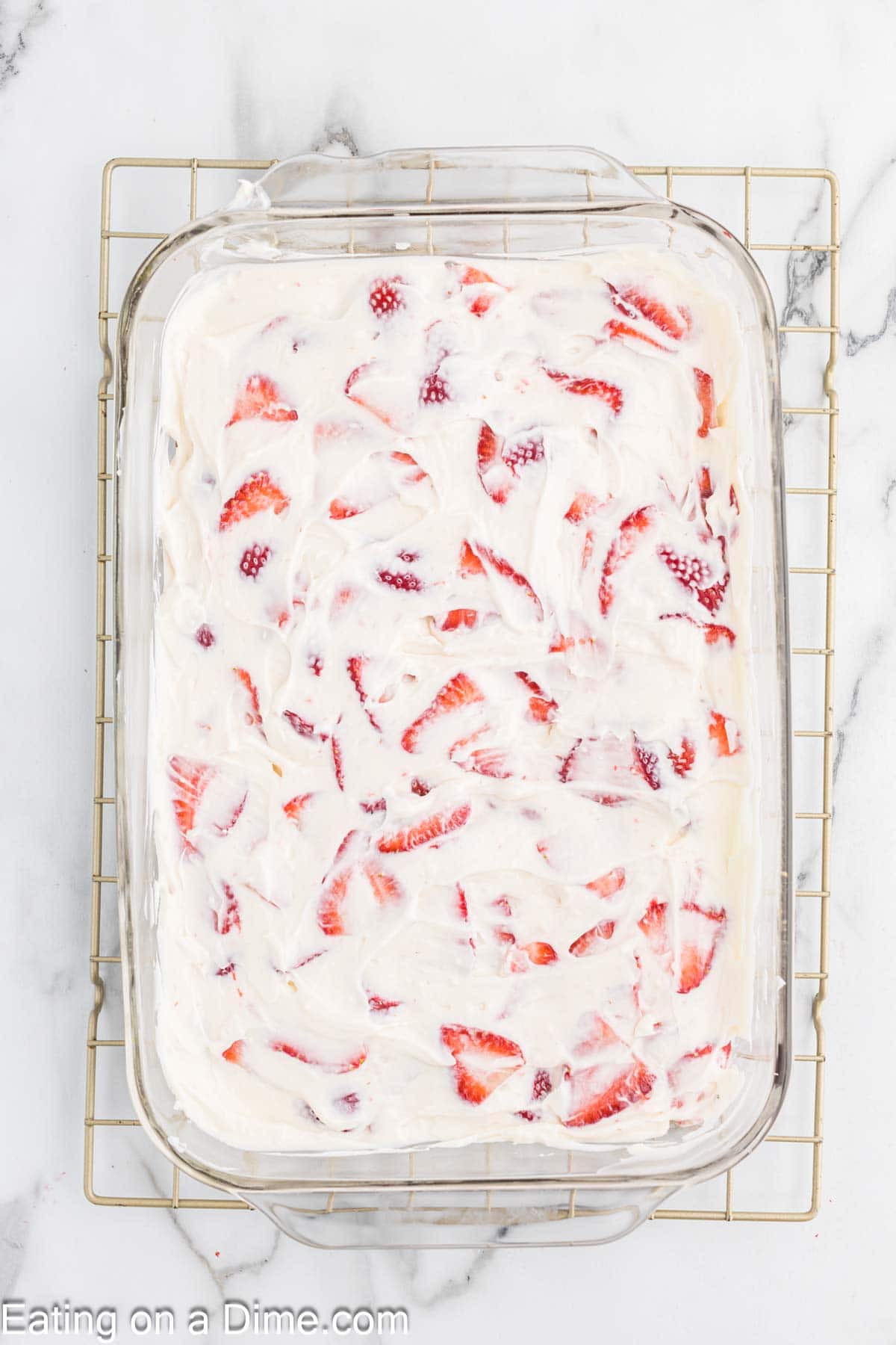 A glass baking dish filled with a white cream mixture topped with slices of fresh strawberries, placed on a cooling rack against a marble background. The cream layer covers most of the strawberries, leaving some partially visible, reminiscent of a delightful strawberry crunch bars recipe.