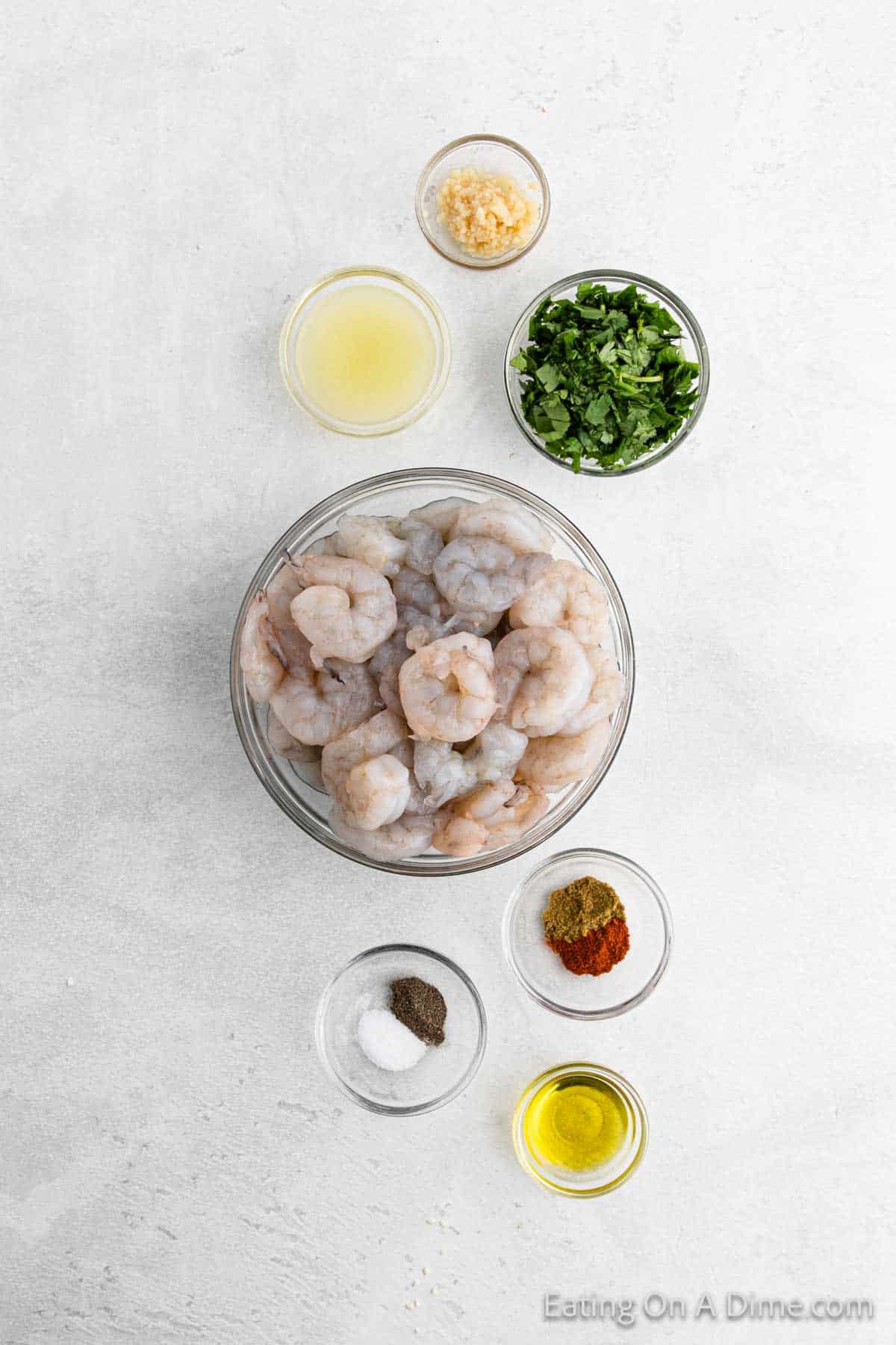 A top-down view of ingredients for Cilantro Lime Shrimp preparation: a bowl of raw shrimp, bowls containing minced garlic, lemon juice, chopped cilantro, and olive oil, along with small dishes of salt, pepper, and spices arranged on a light surface.