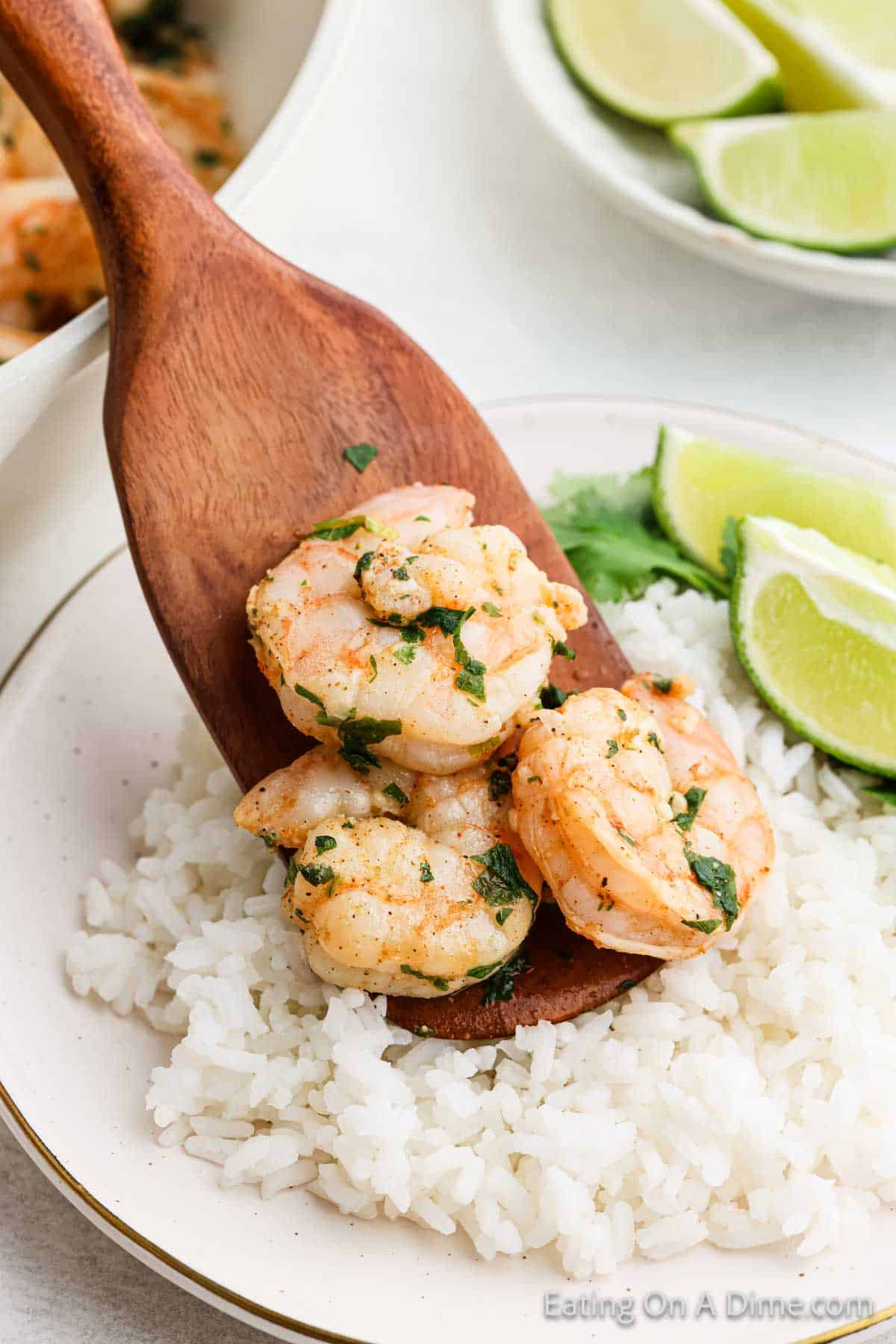 A wooden spoon holds cilantro lime shrimp on a plate of white rice, garnished with fresh cilantro. Slices of lime and cilantro leaves are on the side.