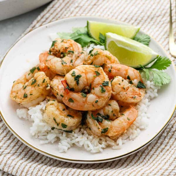 A plate of cilantro lime shrimp garnished with herbs rests on a bed of white rice, accompanied by lime wedges and fresh cilantro leaves. The dish is elegantly set on a striped cloth.