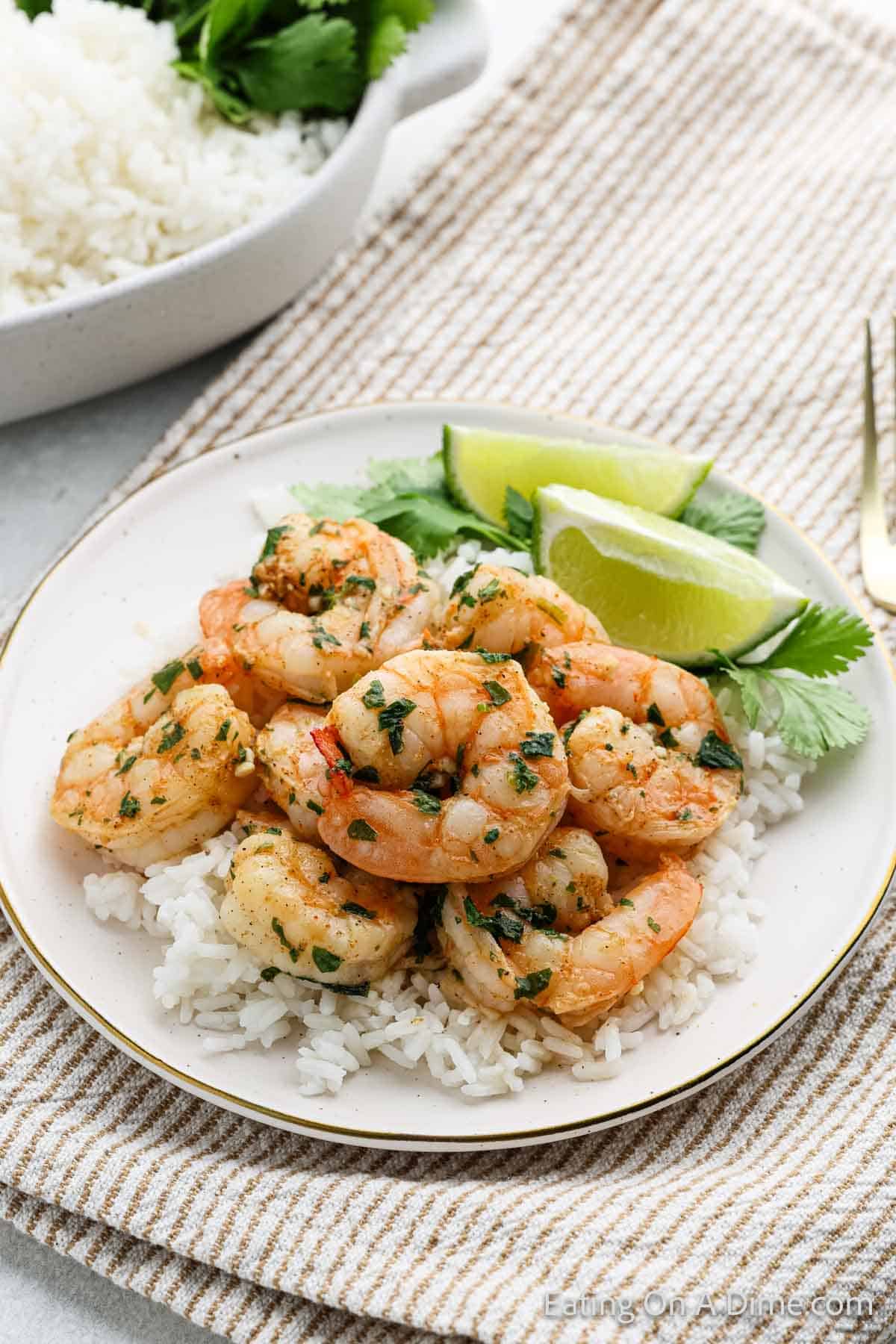 A plate of Cilantro Lime Shrimp garnished with herbs, served over white rice with lime wedges on the side. A dish of rice and fresh cilantro are in the background on a textured cloth.