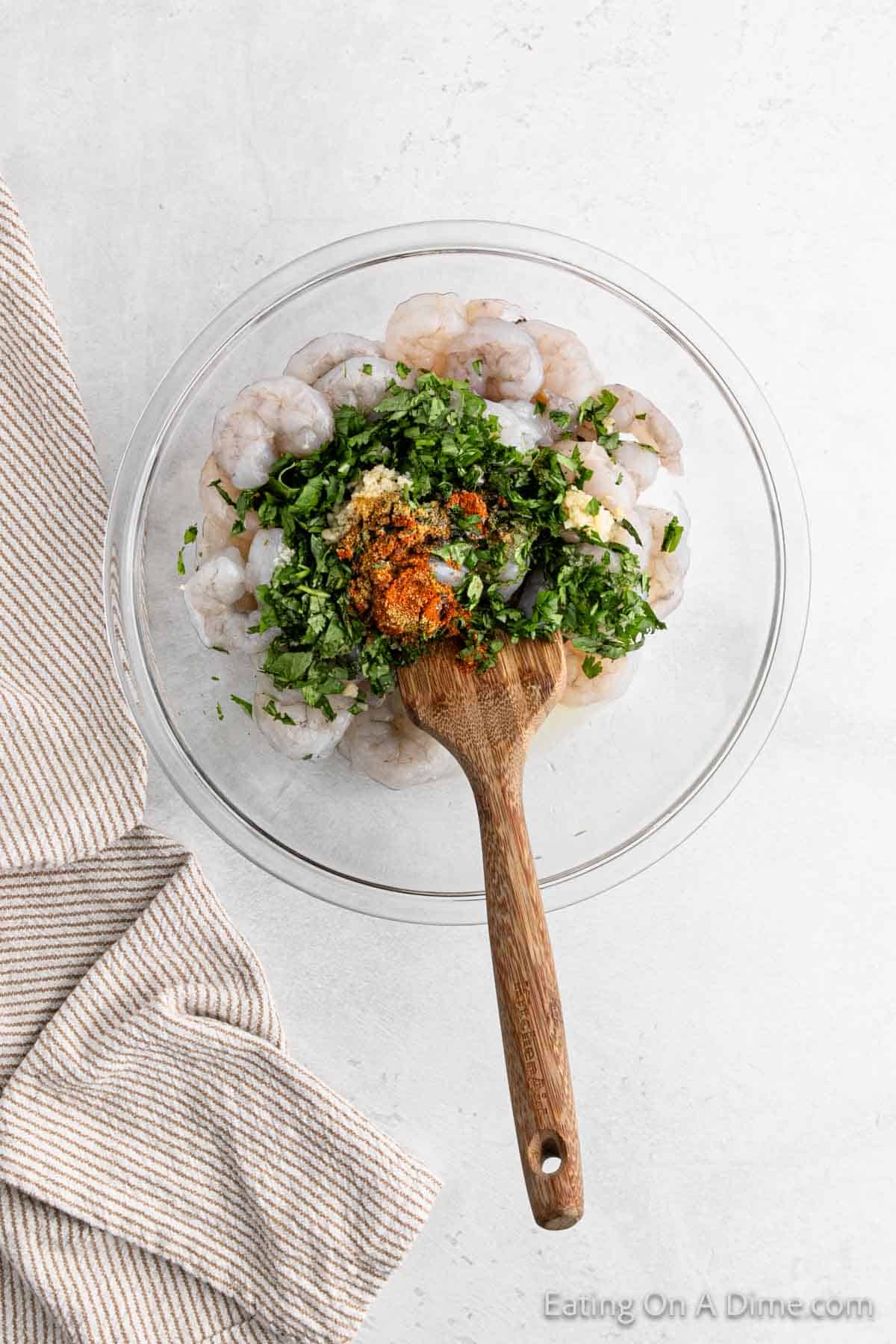 A bowl of Cilantro Lime Shrimp topped with minced garlic and ground spices sits invitingly, the zesty aroma wafting up. A wooden spoon rests in the bowl, ready for serving. Nearby, a striped kitchen towel adds a touch of charm to the light surface.