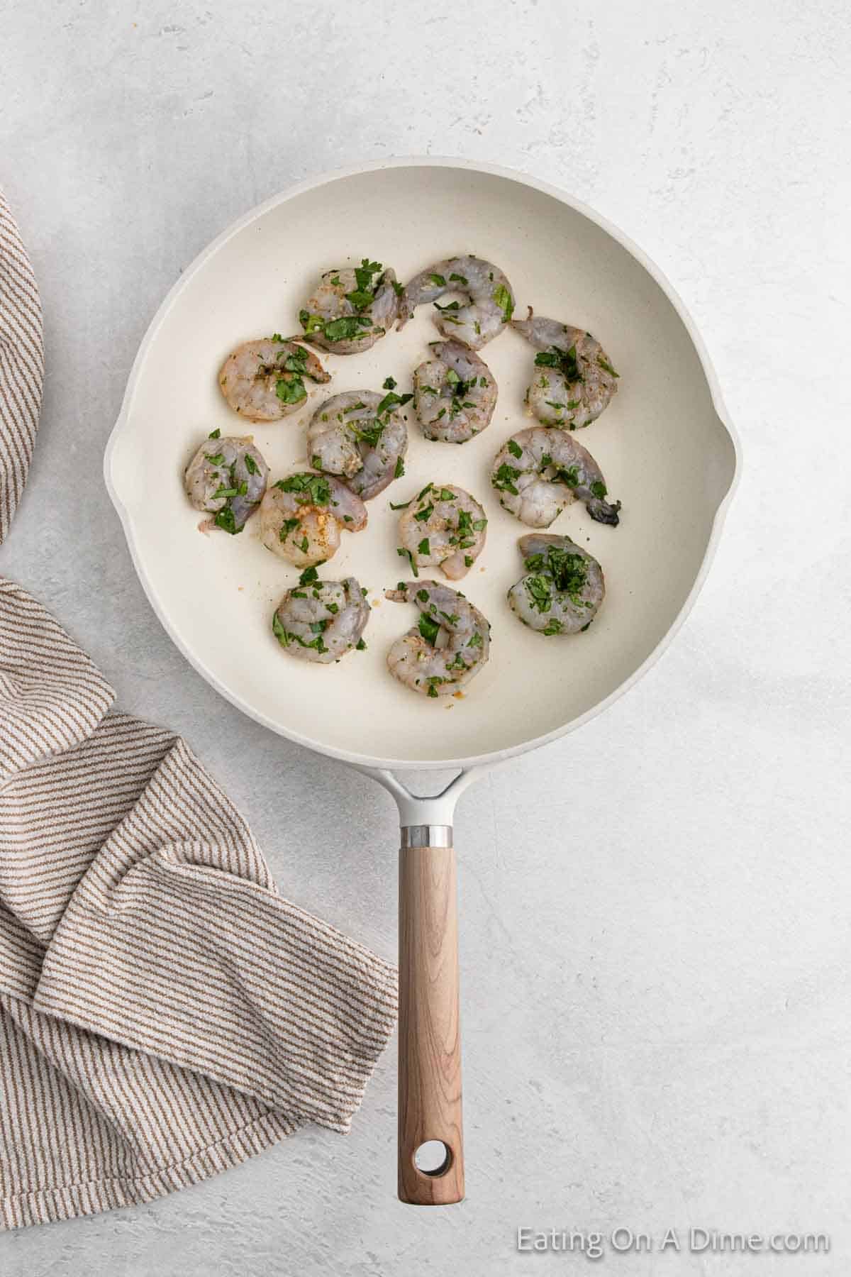 A frying pan with 13 cilantro lime shrimp, sprinkled with chopped herbs, placed on a light-colored surface. A striped cloth lies beside the pan.