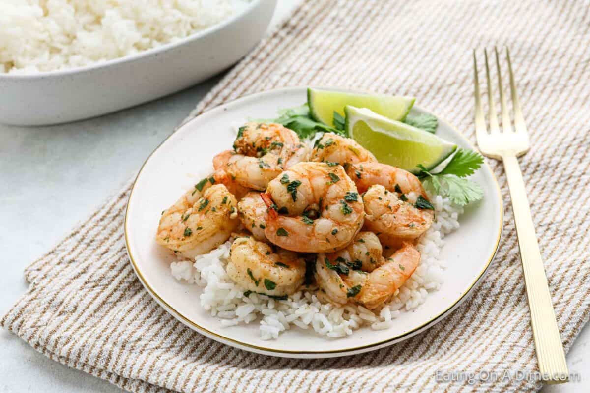 A plate of cilantro lime shrimp sits atop white rice, garnished with fresh cilantro and lime wedges. A gold fork is poised elegantly on a striped cloth, while a bowl of rice waits in the background.