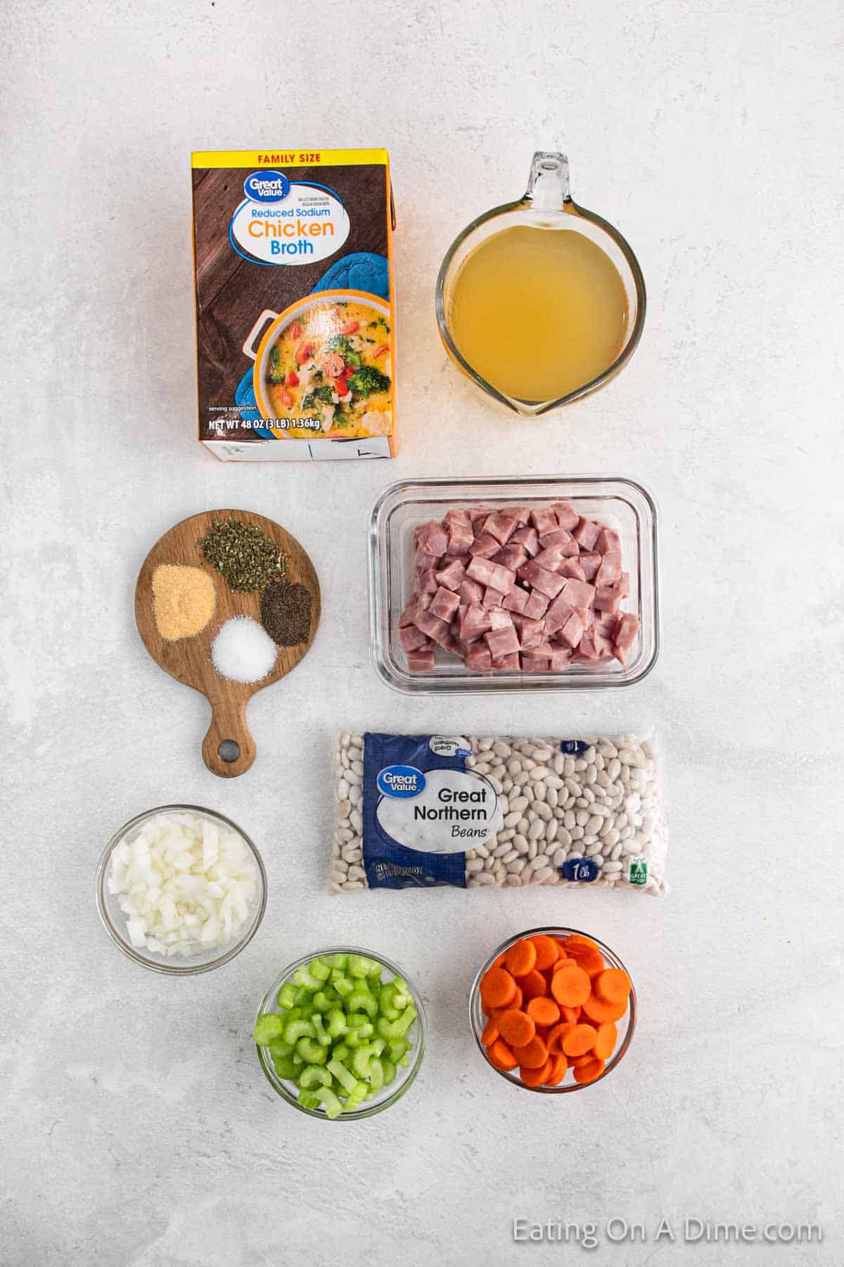 All the essential ingredients for a delightful ham and bean soup crock pot recipe are artfully arranged: a box of chicken broth, a measuring cup with liquid, diced ham in a bowl, Great Northern beans, chopped onions, celery, carrots, and spices on a small board.