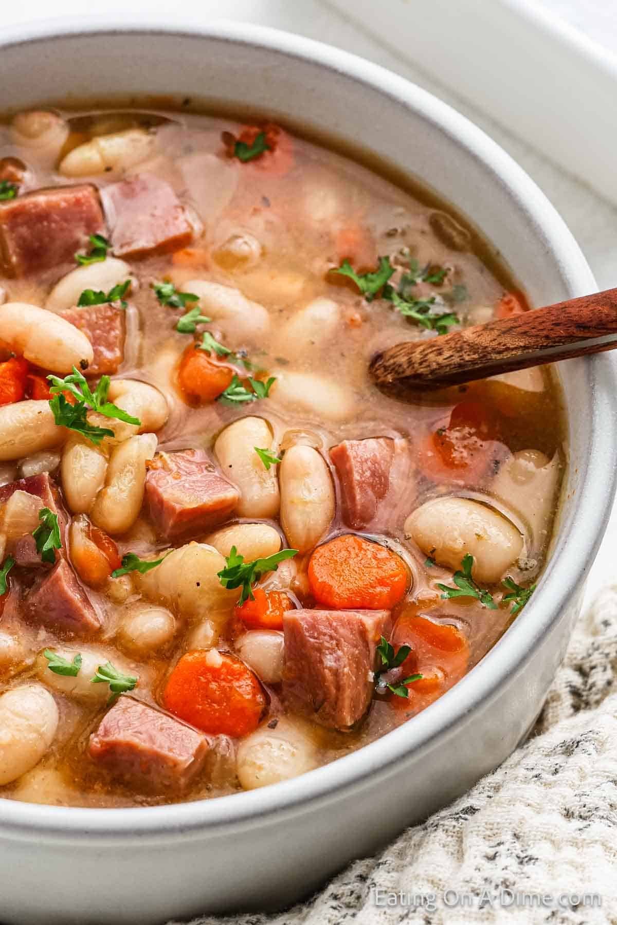 A close-up of a bowl of hearty ham and bean soup reveals tender chunks of ham, white beans, and sliced carrots. Garnished with fresh parsley, this crock pot recipe is complete with a wooden spoon resting inside the bowl on a textured white cloth.