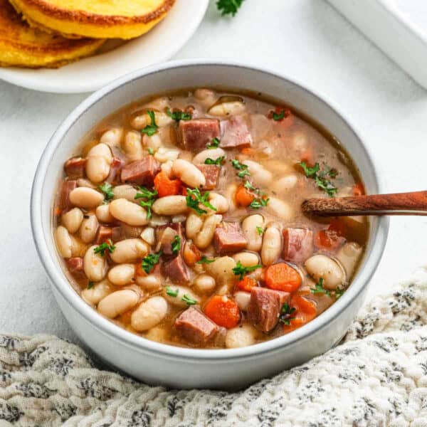 A bowl of hearty white bean and ham soup, a perfect crock pot recipe, is garnished with fresh herbs. Visible ingredients include chunks of ham, carrots, and beans. A wooden spoon rests in the bowl, next to a plate of cornbread on the beige, textured tablecloth.