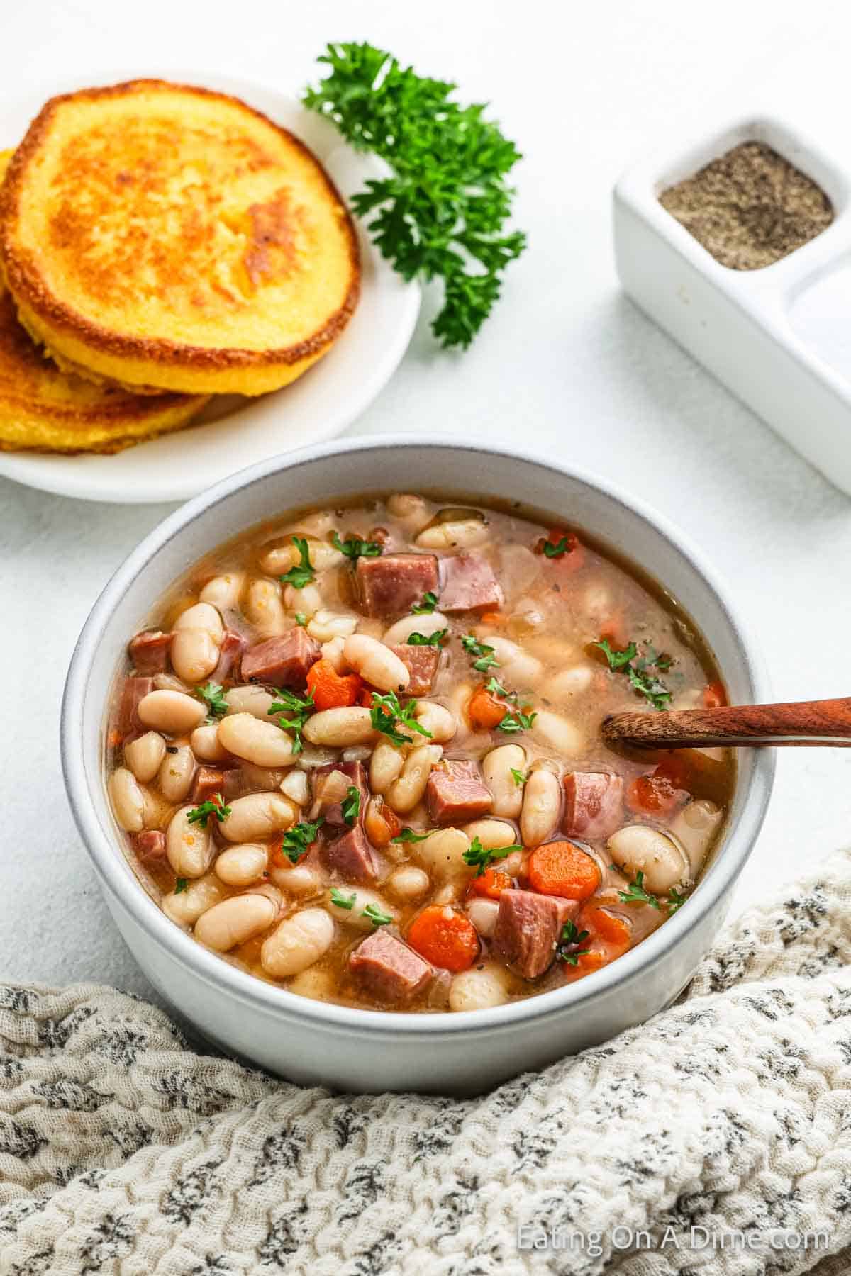 A bowl of hearty ham and bean soup, made with a crock pot recipe, features chunks of carrots and herbs. It's placed next to a stack of cornbread and a small dish of ground pepper. A sprig of parsley completes the cozy scene.