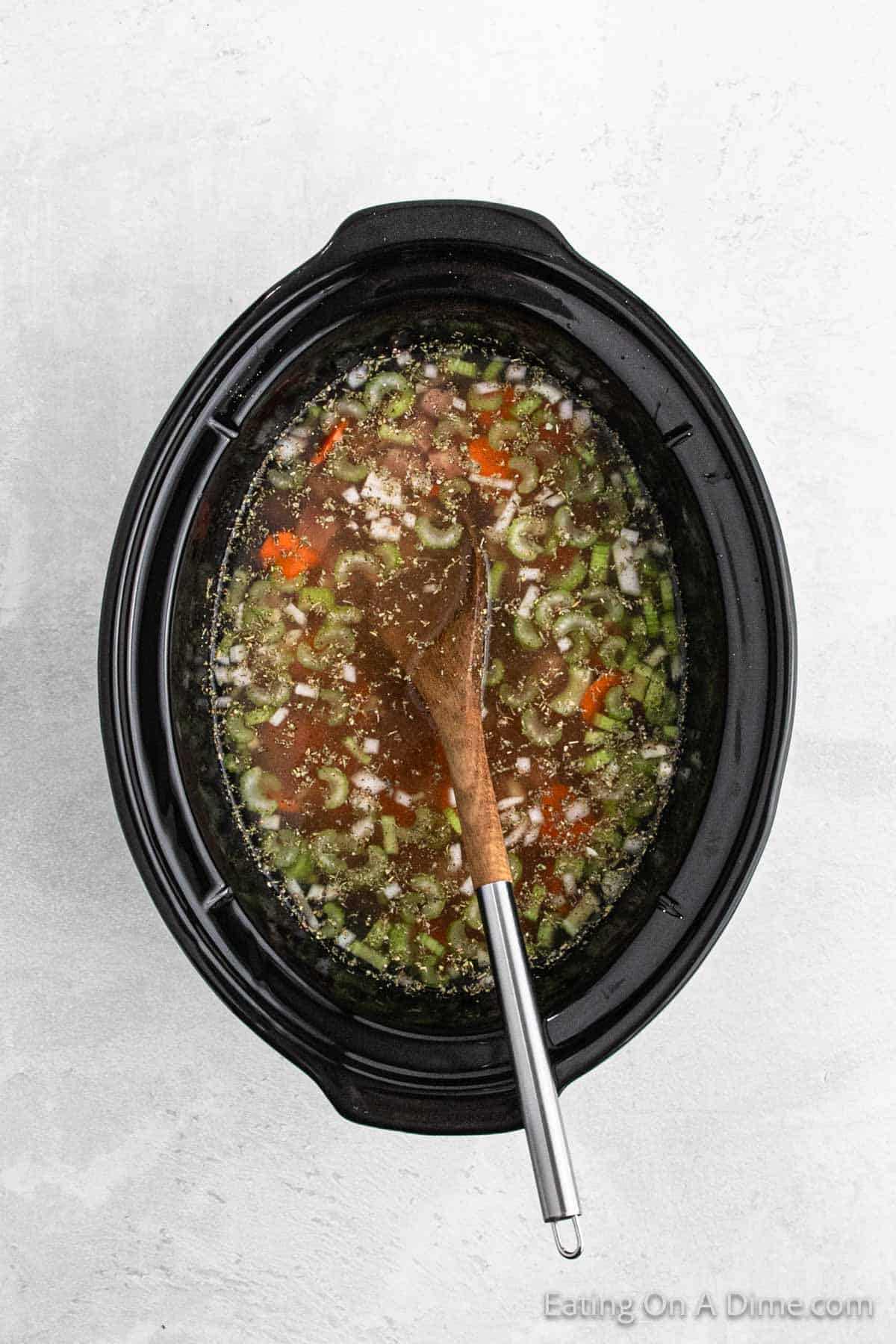 A slow cooker filled with a colorful ham and bean soup, including carrots, celery, and herbs. A wooden spoon rests inside the crock pot, gently stirring the soup's hearty ingredients.