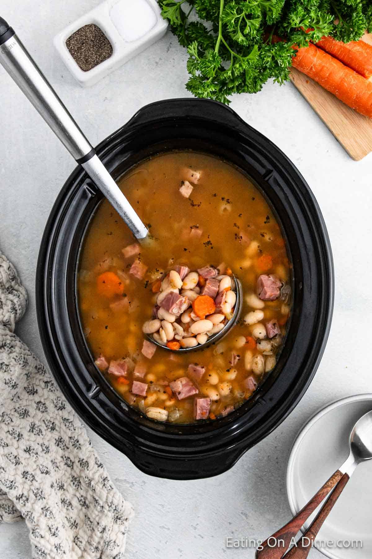 A slow cooker, brimming with a delicious ham and bean soup recipe, features a ladle serving a portion. Ingredients like carrots are visible. Fresh parsley and carrots sit on a cutting board nearby, while a fabric napkin lies beside this classic crock pot dish.