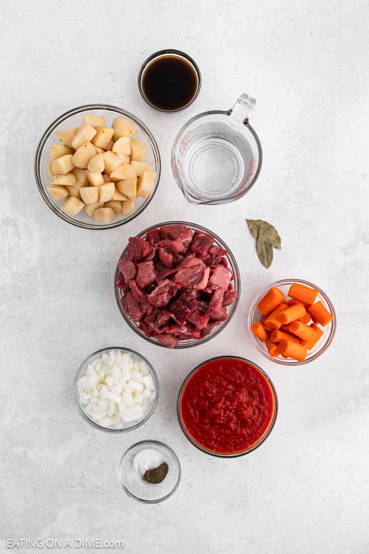 An array of ingredients artfully spread across a light surface includes diced potatoes, diced beef, chopped carrots, diced onions, seasonings, a measuring cup of water, soy sauce, bay leaves, and crushed tomatoes—perfect for creating a savory Instant Pot Beef Stew Recipe.