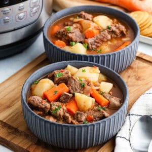 Two gray bowls brimming with Instant Pot beef stew, featuring chunks of beef, carrots, and potatoes, garnished with herbs. The bowls rest on a wooden tray alongside crackers and a spoon, with the trusty pressure cooker visible in the background.