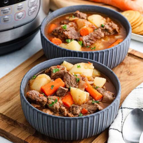 Two gray bowls brimming with Instant Pot beef stew, featuring chunks of beef, carrots, and potatoes, garnished with herbs. The bowls rest on a wooden tray alongside crackers and a spoon, with the trusty pressure cooker visible in the background.