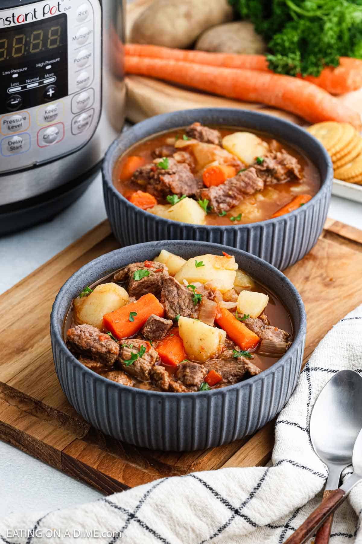 Two bowls of beef stew, part of our Instant Pot Beef Stew Recipe, are filled with tender beef, carrots, and potatoes, garnished with parsley on a wooden board. A cloth napkin and spoon sit beside them, while an Instant Pot, raw carrots, and greens linger in the background.