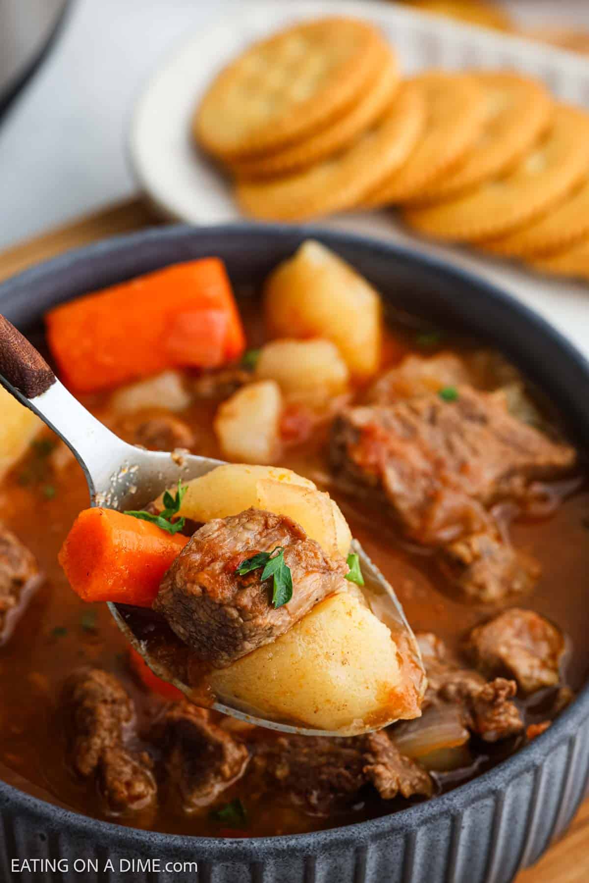 A spoonful of Instant Pot beef stew, brimming with tender chunks of beef, potatoes, and carrots, is being lifted from a bowl. In the background, a plate holds a stack of round crackers.