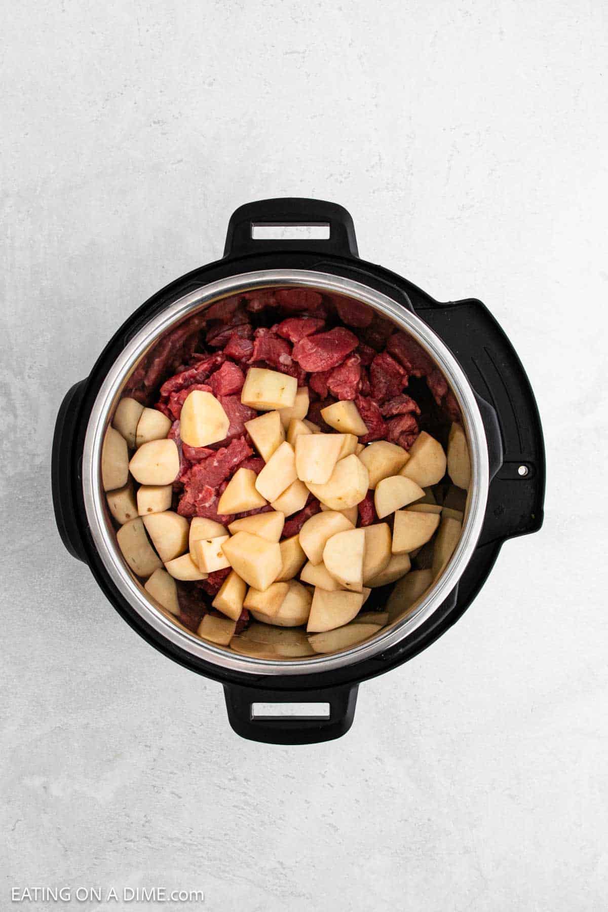 An overhead view of an instant pot reveals uncooked beef chunks and potato pieces, hinting at a delicious Instant Pot Beef Stew Recipe, all set against a light gray background.