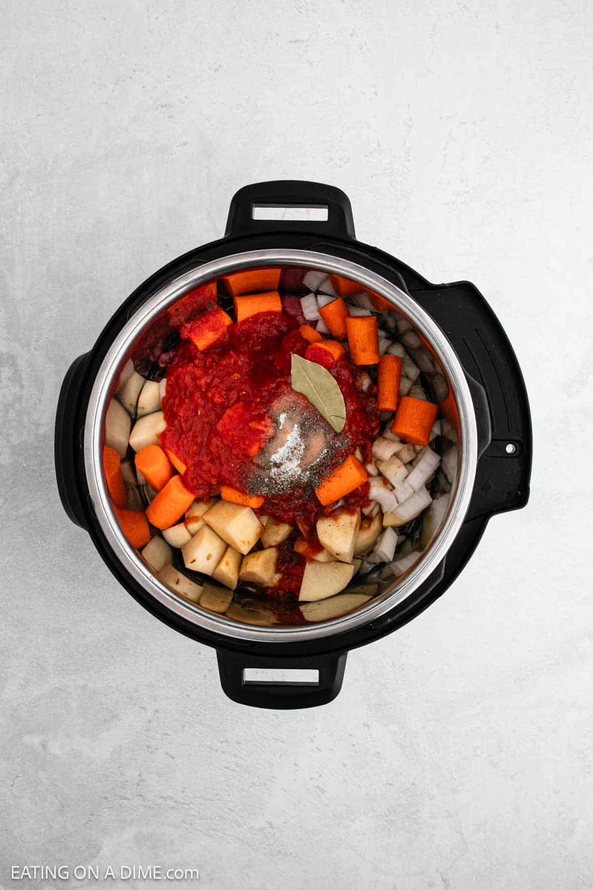 Top view of an open black pressure cooker filled with a colorful mix of chopped vegetables including potatoes, carrots, and onions, topped with tomatoes, spices, and a bay leaf—perfect for an Instant Pot beef stew recipe on a sleek light gray countertop.