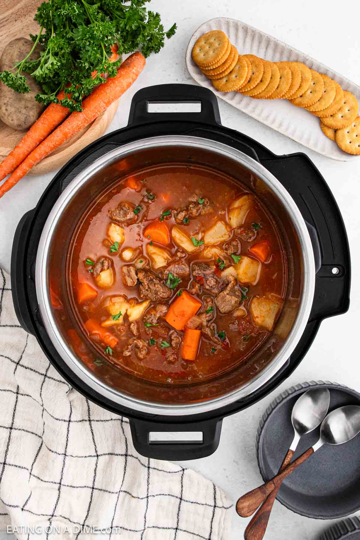 A top view of an Instant Pot Beef Stew recipe features tender chunks of beef, potatoes, and carrots. Fresh parsley and carrots sit nearby, while a plate with crackers and two spoons rests on a checkered cloth beside the pot.