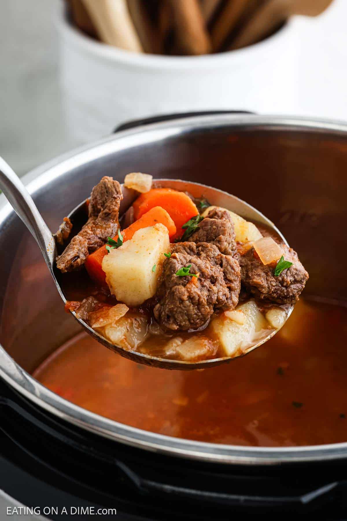 A ladle filled with Instant Pot beef stew, featuring chunks of beef, potatoes, carrots, and herbs, is held above a pot of soup. The rich reddish-brown broth is visible. In the background, a blurred kitchen utensil holder adds to the cozy cooking scene.