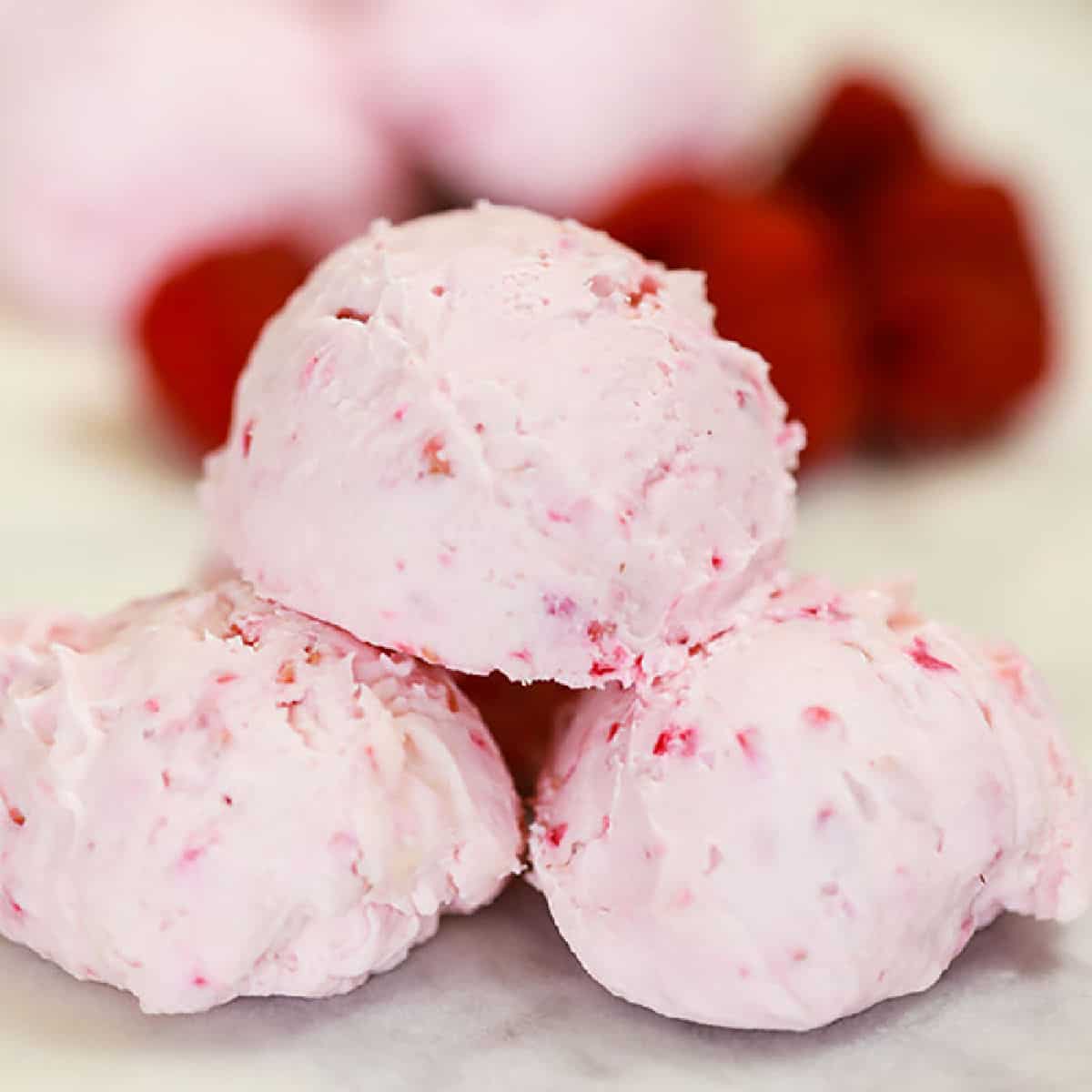 Three scoops of pink raspberry ice cream, reminiscent of a keto cheesecake fat bomb, are stacked on a white surface, with blurred red raspberries in the background. The ice cream has a creamy texture with visible specks of raspberry.