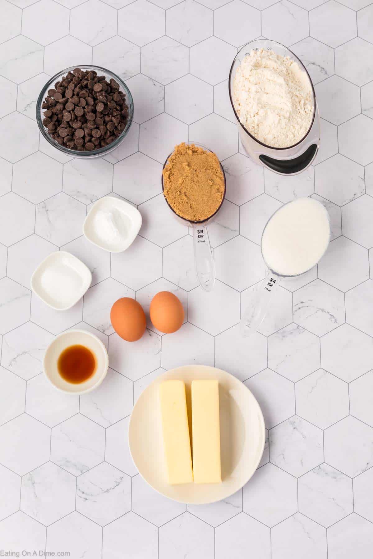 Ingredients for baking a giant cookie recipe are artfully arranged on a hexagonal tile background: chocolate chips, flour, brown sugar, white sugar, baking powder, salt, two eggs, vanilla extract, and two sticks of butter.