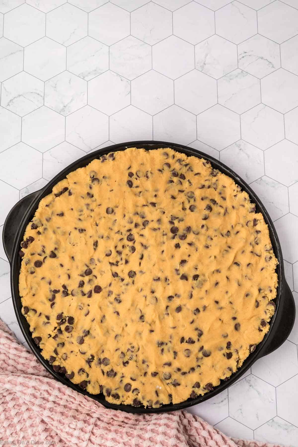 A giant cookie recipe comes to life with unbaked chocolate chip dough spread in a round pan on a geometric-patterned countertop. A pink checkered cloth peeks from the bottom left, adding a touch of charm.