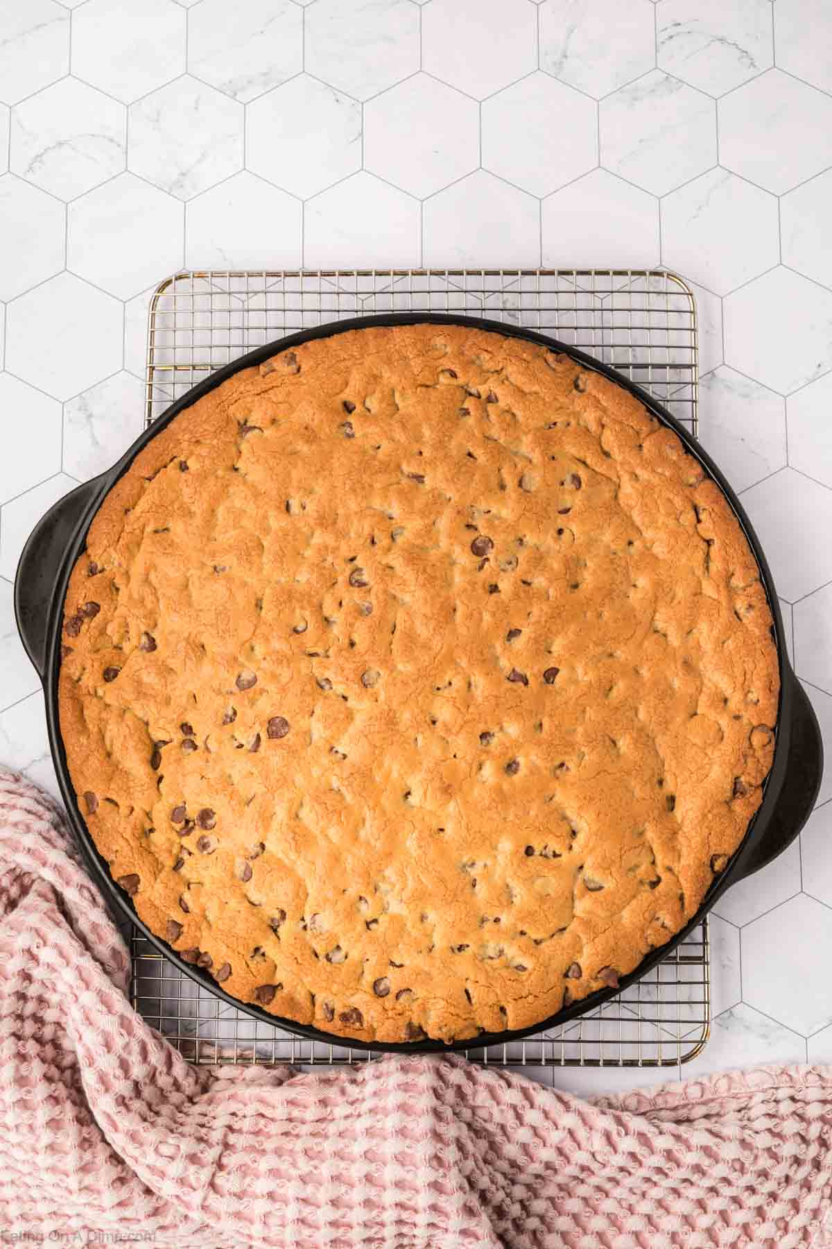 A giant cookie recipe comes to life as a large, freshly baked chocolate chip cookie rests on a round black baking tray atop a cooling rack. A pink and white checkered cloth is partially wrapped around the tray, set against a background of light hexagonal tile design.