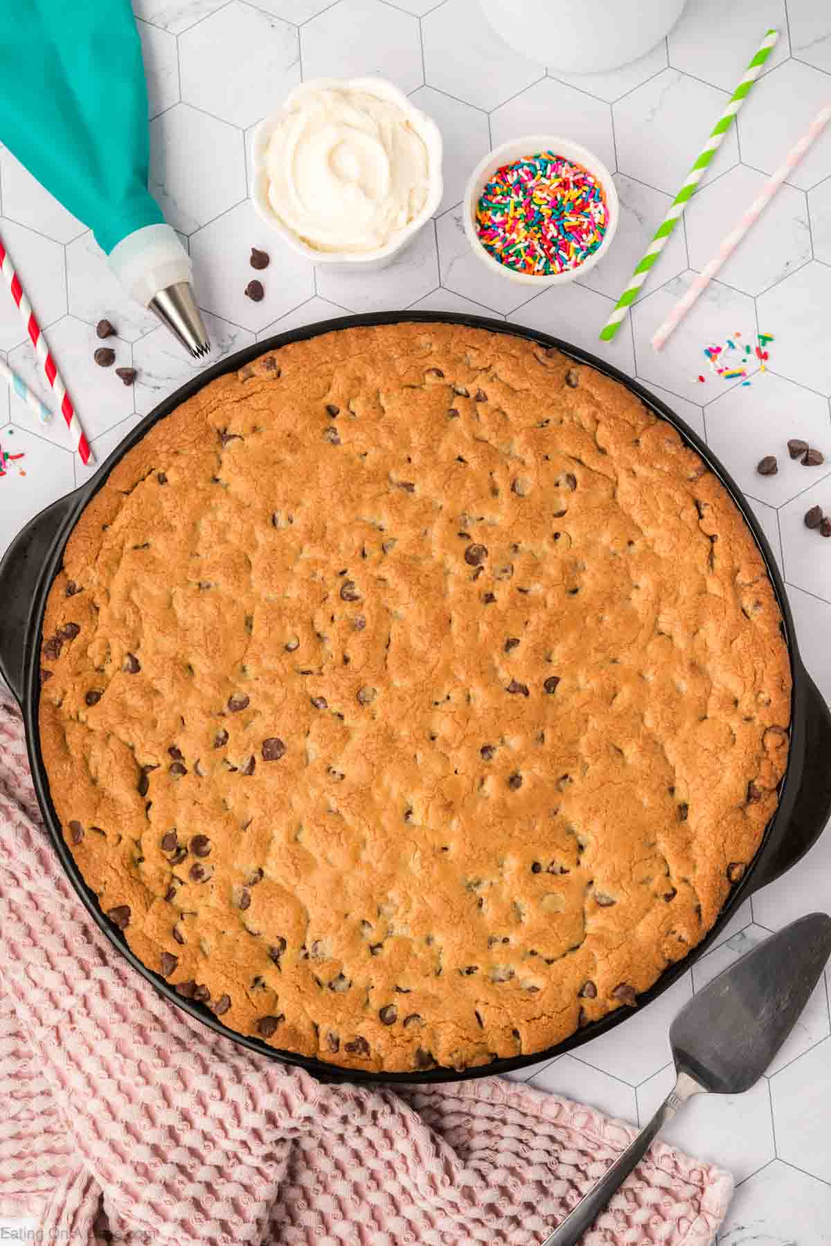 A giant cookie recipe comes to life as a large chocolate chip cookie in a skillet, surrounded by colorful sprinkles, frosting, and straws on a hexagonal patterned surface. Nearby, a piping bag and pink checkered cloth add charm, while a cake server waits on the right.