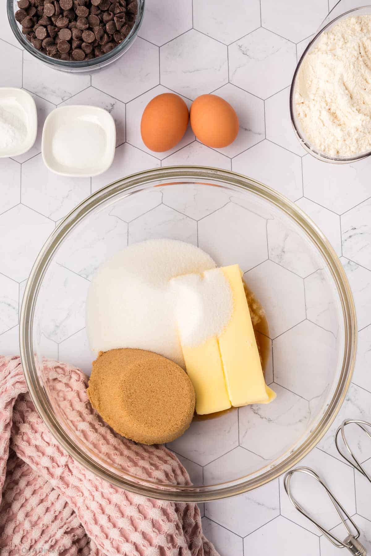 A glass bowl with butter, brown sugar, granulated sugar, and vanilla extract sits ready for a giant cookie recipe. Nearby are eggs, flour, chocolate chips, salt, baking soda, a pink textured cloth, and a whisk on a hexagonal patterned surface.