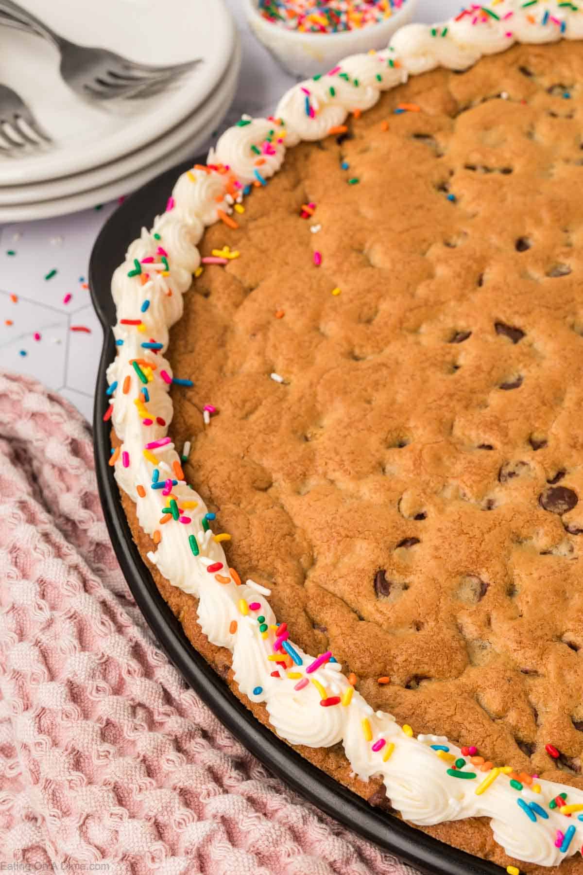 A large chocolate chip cookie cake, crafted from a giant cookie recipe, boasts a border of white frosting and colorful sprinkles on the table. Plates, forks, and a pink textured cloth are nearby. In the background, a bowl brims with extra sprinkles.