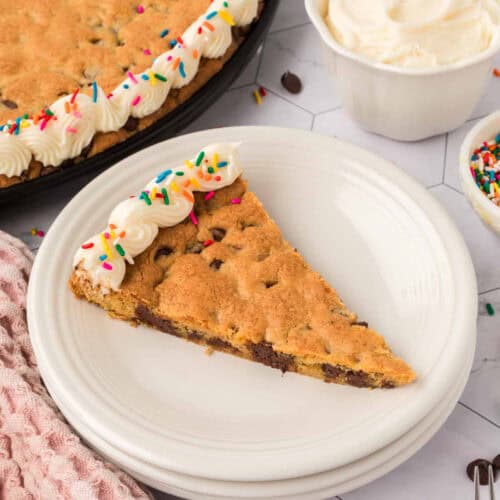 A slice of cookie cake with chocolate chips on a white plate, crafted from a giant cookie recipe. The cake is topped with piped white frosting and colorful sprinkles. Nearby, there's a bowl of frosting and a small dish of sprinkles for extra decoration.