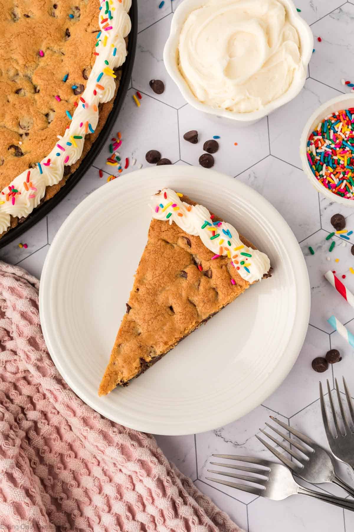 A slice of giant chocolate chip cookie cake with frosting and rainbow sprinkles sits on a white plate. Nearby, the whole cookie cake, a bowl of frosting, and sprinkles rest on a hexagonal marble surface. A pink textured cloth and forks complete this sweet scene.