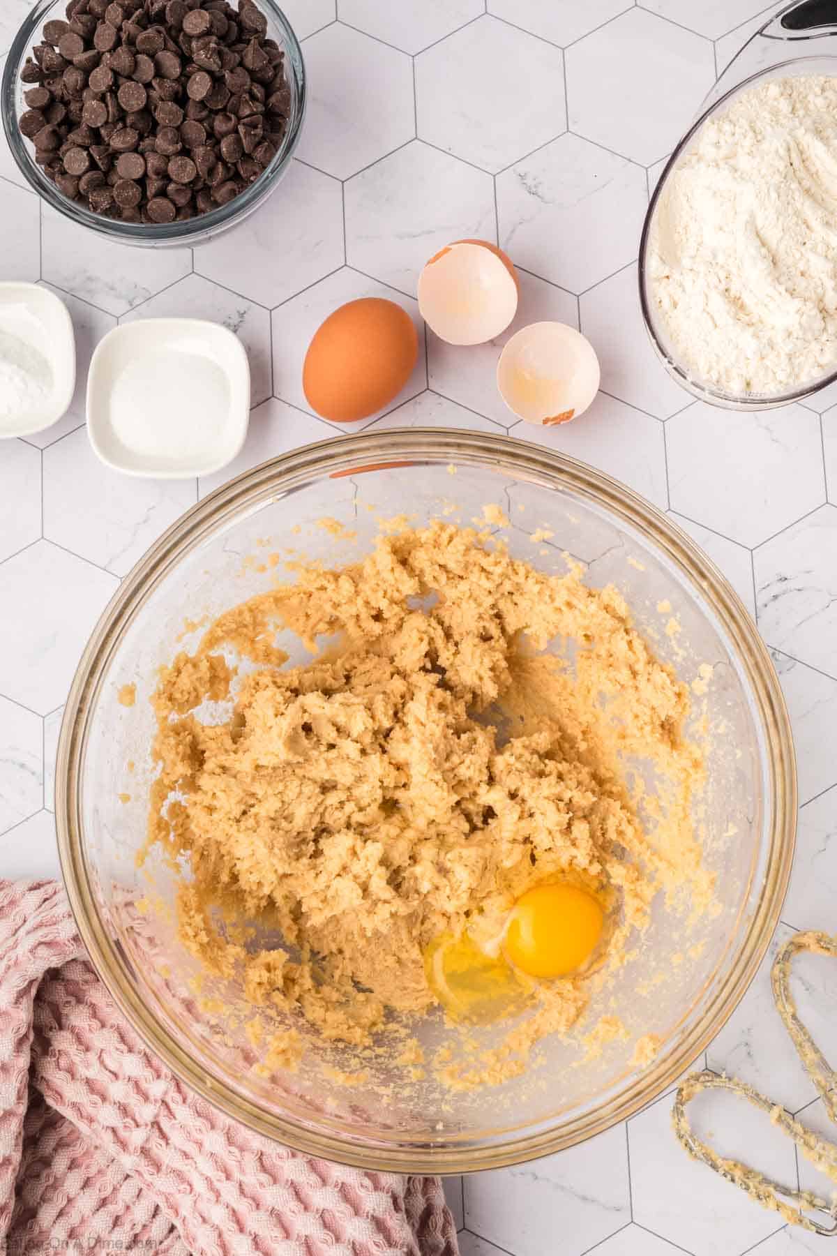 A bowl brimming with cookie dough mixture, ready for a giant cookie recipe, sits at the center with an egg nestled in the middle. Surrounding it are chocolate chips, cracked eggs, and flour on a hexagon-patterned countertop. Nearby, a pink cloth adds a touch of charm.