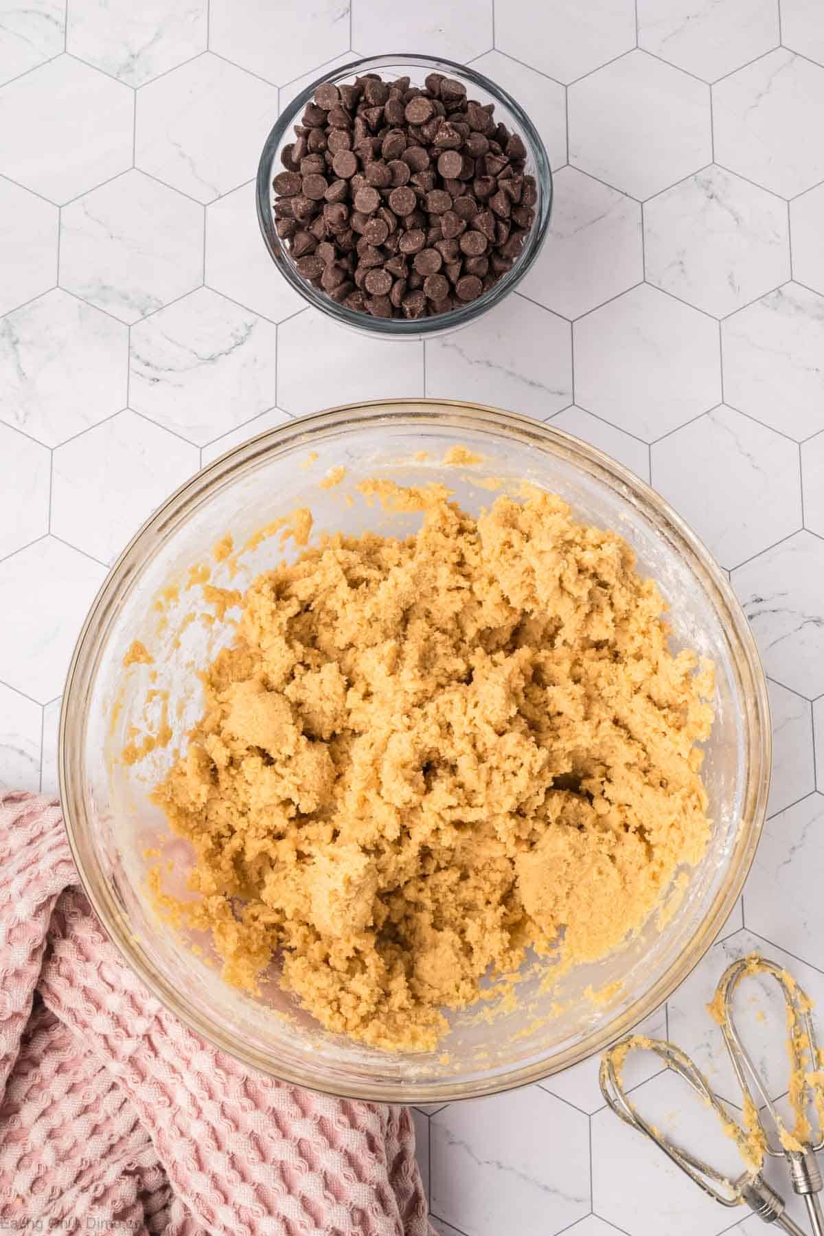 A glass bowl filled with cookie dough for a giant cookie recipe sits on a hexagonal tile surface. Nearby, there's a smaller bowl of chocolate chips. An electric mixer with dough residue and a pink textured cloth are also visible.